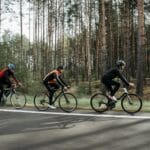 Grupa kolarzy jedzie szosą w słońcu, w tle zielony las