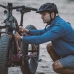 Mężczyzna w kasku naprawia rower elektryczny na piaszczystym szlaku